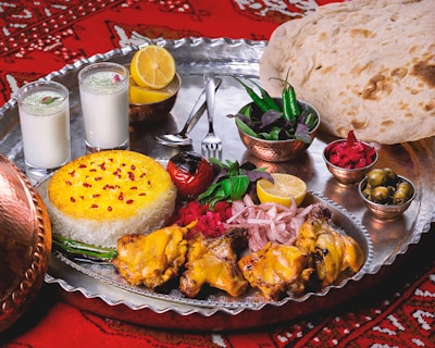 A lavish spread of Middle Eastern or South Asian cuisine is presented on a large metal tray. There are servings of yellow saffron rice and grilled chicken with a side of onions, garnished with lemon and fresh herbs. Two glasses of a yogurt-based drink, possibly lassi, are seen alongside. Additional side dishes include a bowl of green chili peppers and herbs, pickled vegetables, and a small bowl of olives. A large flatbread rests on the side, and the scene is set against a red patterned fabric.