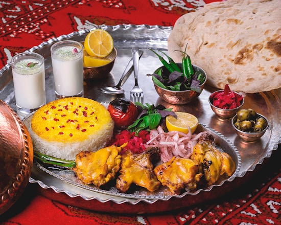 A lavish spread of Middle Eastern or South Asian cuisine is presented on a large metal tray. There are servings of yellow saffron rice and grilled chicken with a side of onions, garnished with lemon and fresh herbs. Two glasses of a yogurt-based drink, possibly lassi, are seen alongside. Additional side dishes include a bowl of green chili peppers and herbs, pickled vegetables, and a small bowl of olives. A large flatbread rests on the side, and the scene is set against a red patterned fabric.