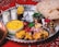 A lavish spread of Middle Eastern or South Asian cuisine is presented on a large metal tray. There are servings of yellow saffron rice and grilled chicken with a side of onions, garnished with lemon and fresh herbs. Two glasses of a yogurt-based drink, possibly lassi, are seen alongside. Additional side dishes include a bowl of green chili peppers and herbs, pickled vegetables, and a small bowl of olives. A large flatbread rests on the side, and the scene is set against a red patterned fabric.