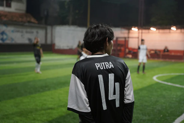 A person wearing a black and white soccer jersey with the name 'Putra' and the number '14' on the back is standing on a soccer field. The field is made of artificial turf and is illuminated by lights. Additional players are blurred in the background, suggesting an ongoing soccer match or practice.