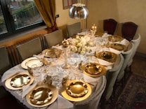 A dining table is elegantly set with a white tablecloth, gold plates, and gold accents. There are napkins folded neatly, surrounded by crystal glasses, candles, and a floral centerpiece with white and cream flowers. The setting includes plush chairs and a warm, ambient lighting from a pendant lamp above.