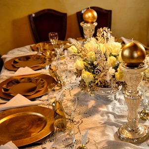 An elegantly set dining table with ornate golden decorative plates, glassware, and silverware arranged on an intricate white lace tablecloth. A luxurious floral centerpiece composed of cream roses and festive gold foliage is positioned in the center. The table is further adorned with golden spherical ornaments placed on tall silver candlesticks, adding a touch of opulence.