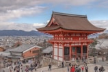 A traditional Japanese shrine or temple with vibrant red and white colors stands prominently, surrounded by a busy scene of numerous visitors milling around the courtyard. The background features a sprawling urban landscape, with distant mountains under a partly cloudy sky.