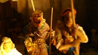 A detailed depiction of intricately crafted figurines made from a material such as clay or wood, with expressive facial features and clothing indicative of historic or cultural attire. The scene is dimly lit, creating dramatic shadows and emphasizing the craftsmanship of the figures.