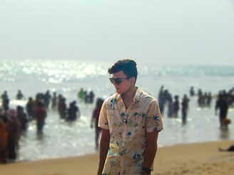 A young person wearing sunglasses and a floral shirt stands on a sandy beach. In the background, there is a busy scene with many people enjoying the water, indicating a lively beach environment. The sun glistens off the ocean, creating a sparkling effect.