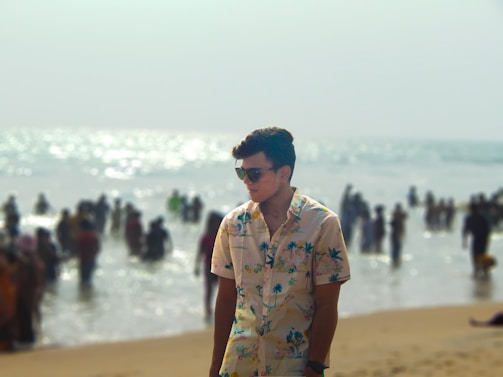 A young person wearing sunglasses and a floral shirt stands on a sandy beach. In the background, there is a busy scene with many people enjoying the water, indicating a lively beach environment. The sun glistens off the ocean, creating a sparkling effect.