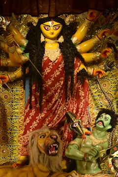 A vibrant and detailed statue of a goddess with multiple arms, adorned in a red saree with gold ornaments, holding various weapons. The figure stands dominantly over a lion beside a green-skinned figure. The background is richly decorated with intricate patterns and vivid colors.