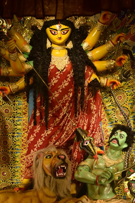 A vibrant and detailed statue of a goddess with multiple arms, adorned in a red saree with gold ornaments, holding various weapons. The figure stands dominantly over a lion beside a green-skinned figure. The background is richly decorated with intricate patterns and vivid colors.