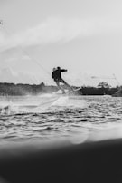 A person engages in wakeboarding on a body of water, creating splashes and waves. The scene appears dynamic and energetic, set against a backdrop of trees and a cloudy sky.