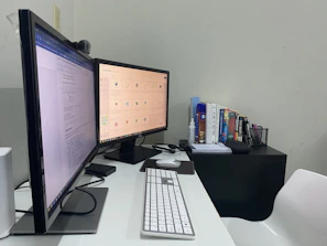 A home office setup featuring two monitors placed side by side, displaying different content. A keyboard and mouse are positioned on a white desk, with a webcam perched on one of the monitors. To the right, a small table holds a stack of books, and nearby are various stationery items such as notebooks, pens, and a blue canister.