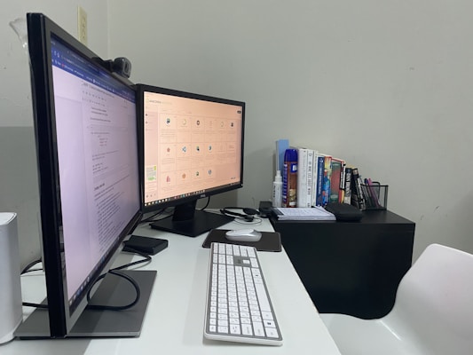 A home office setup featuring two monitors placed side by side, displaying different content. A keyboard and mouse are positioned on a white desk, with a webcam perched on one of the monitors. To the right, a small table holds a stack of books, and nearby are various stationery items such as notebooks, pens, and a blue canister.