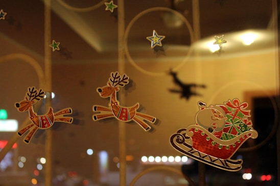 Three reindeer stickers are placed on a glass surface, appearing to pull Santa Claus in a sleigh laden with gifts. Star stickers are scattered around, adding to a festive atmosphere. The background features soft lighting and a blurred shadow of Santa, enhancing the holiday theme.
