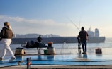 Several people are standing at a waterfront with fishing rods, engaging in fishing activities. A large cargo ship is visible in the background, sailing across the water. The scene is calm with clear skies, and there are various fishing gear items and a bench nearby.