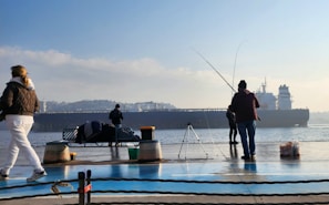 Several people are standing at a waterfront with fishing rods, engaging in fishing activities. A large cargo ship is visible in the background, sailing across the water. The scene is calm with clear skies, and there are various fishing gear items and a bench nearby.