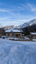 A snow-covered stone house with wooden shutters is set against a backdrop of majestic mountains. The bright blue sky above adds a serene atmosphere to the rustic mountain setting.