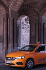 A bright orange taxi is driving through an area with large, intricately designed stone columns and arches. The background shows a stone wall with some greenery growing on it.