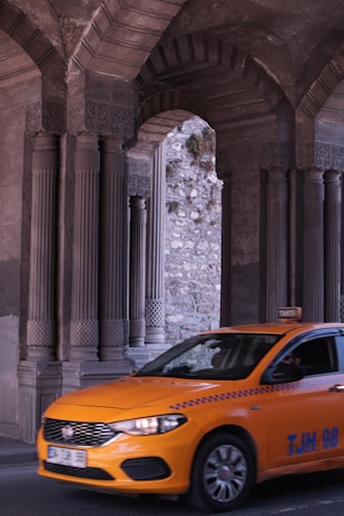A bright orange taxi is driving through an area with large, intricately designed stone columns and arches. The background shows a stone wall with some greenery growing on it.