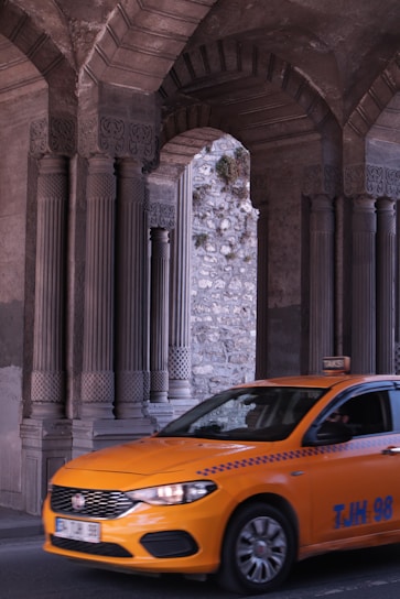 A bright orange taxi is driving through an area with large, intricately designed stone columns and arches. The background shows a stone wall with some greenery growing on it.
