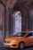A bright orange taxi is driving through an area with large, intricately designed stone columns and arches. The background shows a stone wall with some greenery growing on it.