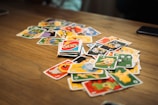 A scattered pile of brightly colored cards with various cartoon characters and numbers is spread out on a wooden table. In the center, an Uno card with distinctive red, blue, yellow, and green design is noticeable.