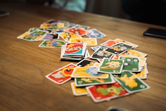 A scattered pile of brightly colored cards with various cartoon characters and numbers is spread out on a wooden table. In the center, an Uno card with distinctive red, blue, yellow, and green design is noticeable.