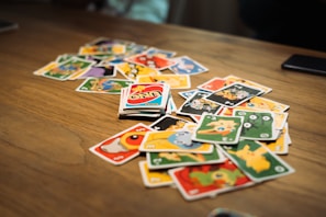A scattered pile of brightly colored cards with various cartoon characters and numbers is spread out on a wooden table. In the center, an Uno card with distinctive red, blue, yellow, and green design is noticeable.