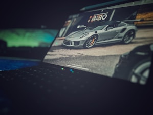 A laptop screen displays an image of a sleek, gray sports car with orange brake calipers, parked on a road. The background shows blurred surroundings, including buildings and greenery. The focus is primarily on the car, with the laptop's keyboard visible in the foreground, slightly out of focus.