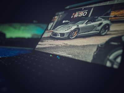 A laptop screen displays an image of a sleek, gray sports car with orange brake calipers, parked on a road. The background shows blurred surroundings, including buildings and greenery. The focus is primarily on the car, with the laptop's keyboard visible in the foreground, slightly out of focus.