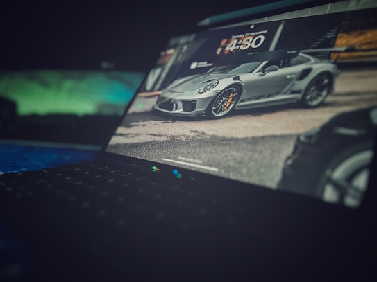 A laptop screen displays an image of a sleek, gray sports car with orange brake calipers, parked on a road. The background shows blurred surroundings, including buildings and greenery. The focus is primarily on the car, with the laptop's keyboard visible in the foreground, slightly out of focus.
