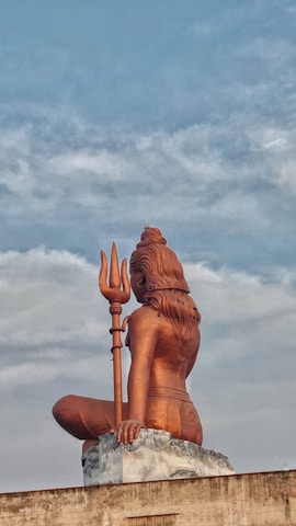 A large, bronze-colored statue of a seated figure holding a trident, viewed from behind. The statue has long hair and sits on a stone pedestal against a backdrop of a cloudy sky.