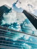 A tall glass skyscraper reflecting the blue sky and fluffy white clouds. The perspective is from the ground looking up, highlighting the building's geometric structure and glossy surface. Two window cleaners are visible on a suspended platform.