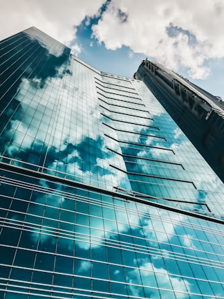 A tall glass skyscraper reflecting the blue sky and fluffy white clouds. The perspective is from the ground looking up, highlighting the building's geometric structure and glossy surface. Two window cleaners are visible on a suspended platform.
