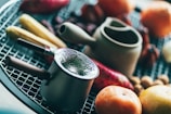 An assortment of cooking utensils and vegetables are placed on a grill. A small metal pot with a textured surface is prominently displayed in the foreground, surrounded by a wooden mortar and pestle. Various vegetables, including carrots and onions, add a splash of color to the scene.