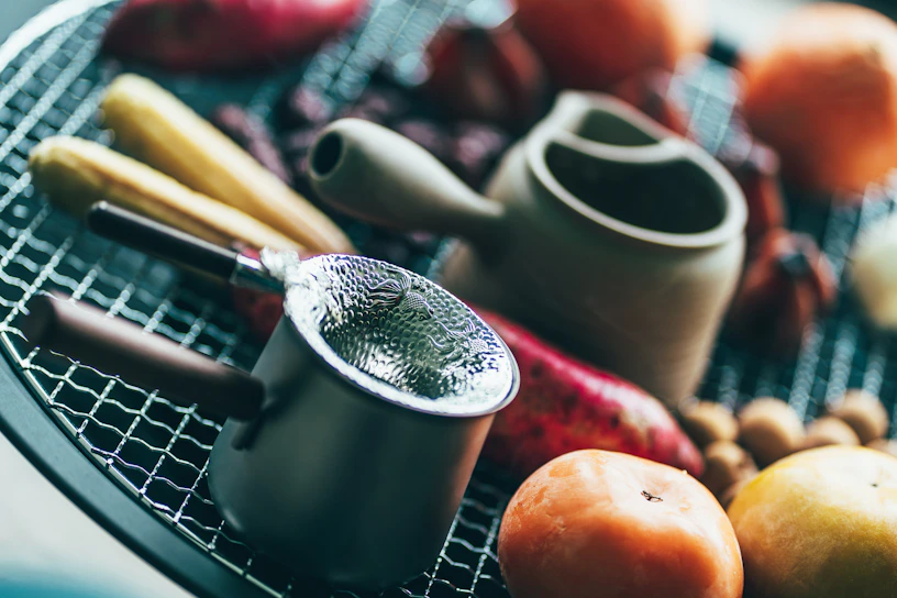 An assortment of cooking utensils and vegetables are placed on a grill. A small metal pot with a textured surface is prominently displayed in the foreground, surrounded by a wooden mortar and pestle. Various vegetables, including carrots and onions, add a splash of color to the scene.