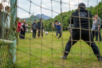 A group of people is gathered on a grassy field, engaged in a casual soccer practice or game. One person is actively kicking a soccer ball towards the goal, while another person stands ready to block in the goalpost. Spectators on both sides appear to be cheering or watching intently. The background features green trees and mountainous landscape visible in the distance.