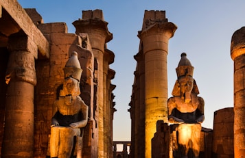 Massive ancient Egyptian statues seated among towering columns, with intricate carvings visible on the stone surfaces. The scene is illuminated by warm artificial lighting against a dusky sky.