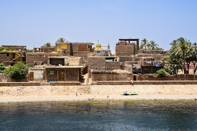 A collection of rustic brick buildings and houses is situated along a riverbank. The structures have flat roofs and some greenery atop them, indicating a mix of rural and residential surroundings. A mosque with a distinct yellow dome stands out among the buildings, surrounded by palm trees and vibrant washing lines. The river in the foreground shows clear, rippling water, with a few people walking along the edge.
