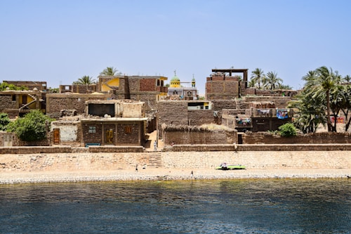 A collection of rustic brick buildings and houses is situated along a riverbank. The structures have flat roofs and some greenery atop them, indicating a mix of rural and residential surroundings. A mosque with a distinct yellow dome stands out among the buildings, surrounded by palm trees and vibrant washing lines. The river in the foreground shows clear, rippling water, with a few people walking along the edge.