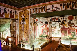 A richly decorated ancient Egyptian tomb with intricate hieroglyphs and colorful wall paintings featuring deities and symbolic figures. The room is illuminated by strategic lighting, showcasing the detail of the artwork and architectural features, including carved pillars and ornate borders.