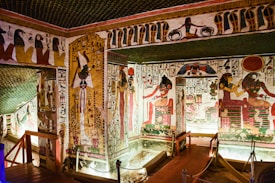 A richly decorated ancient Egyptian tomb with intricate hieroglyphs and colorful wall paintings featuring deities and symbolic figures. The room is illuminated by strategic lighting, showcasing the detail of the artwork and architectural features, including carved pillars and ornate borders.