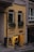 A small storefront with a yellow exterior, featuring two narrow windows on the upper level with wrought iron bars and potted plants. Below, the entrance to the store displays various leather goods, highlighted by warm lighting. The sign outside reads 'MORIA # Leathergoods' and there is a wooden plaque indicating 'Since 2012'.