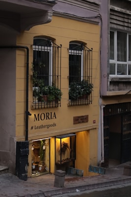 A small storefront with a yellow exterior, featuring two narrow windows on the upper level with wrought iron bars and potted plants. Below, the entrance to the store displays various leather goods, highlighted by warm lighting. The sign outside reads 'MORIA # Leathergoods' and there is a wooden plaque indicating 'Since 2012'.