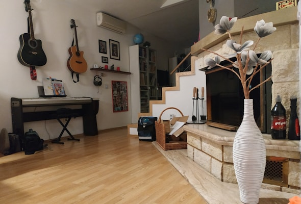 A cozy room with a wooden floor featuring a digital piano on the left and two guitars hanging on the wall above it. There is a shelf with picture frames and small decorative items. A stone fireplace is in the corner, adorned with a large white vase holding decorative branches. Bookshelves are visible in the background near a staircase.