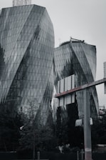 Two modern high-rise buildings with a unique, curving architectural design are featured. The structures are covered in glass panels which reflect the surrounding environment. At the base, there is dense greenery with trees and plants. In the foreground, parts of a billboard structure and a sign are visible.