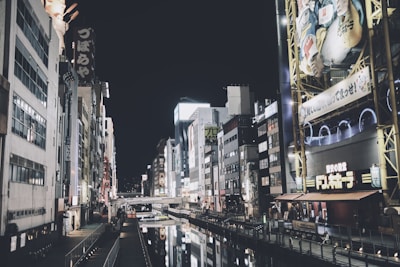 A nighttime urban scene featuring a canal lined with tall buildings adorned with bright neon signs and advertisements. The water in the canal reflects the illuminated signs and structures, creating a vibrant ambiance. The street lights and signage create a mixed lighting environment with various light sources.