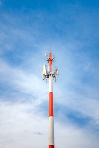 A tall communication tower with a vibrant red and white color scheme stands against a clear, bright blue sky. The structure features multiple antennae and equipment mounted towards the top.