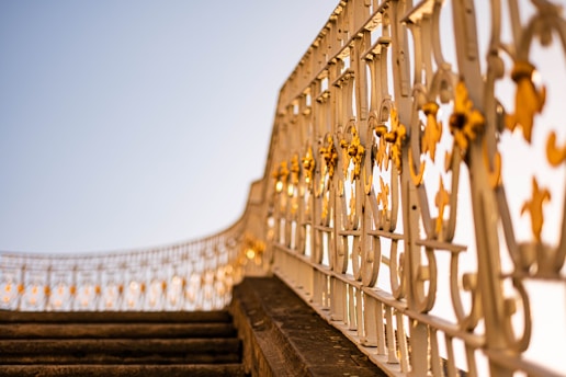 A decorative wrought iron railing with intricate designs and golden accents, extending along a stairway. The image is taken from a low angle, giving prominence to the artistic metalwork against a clear blue sky.