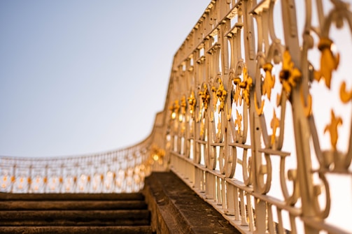 A decorative wrought iron railing with intricate designs and golden accents, extending along a stairway. The image is taken from a low angle, giving prominence to the artistic metalwork against a clear blue sky.