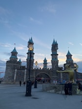 A fantasy castle structure with tall stone towers and teal conical rooftops standing against a dusk sky. In the forefront, there is a large golden statue of a whimsical frog with an open mouth and outstretched arms. Nearby, a few people are gathered on a paved area with ornate street lamps and fencing.