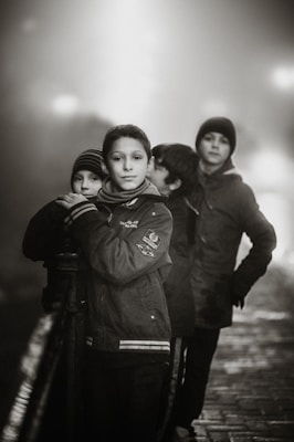 Four boys stand together in a foggy, outdoor setting. They are wearing winter clothing, including jackets and beanies. The scene is captured in black and white, adding a timeless and somber feel. The boys are leaning against a railing on what appears to be a bridge or pathway, with cobblestone-like textures underfoot.